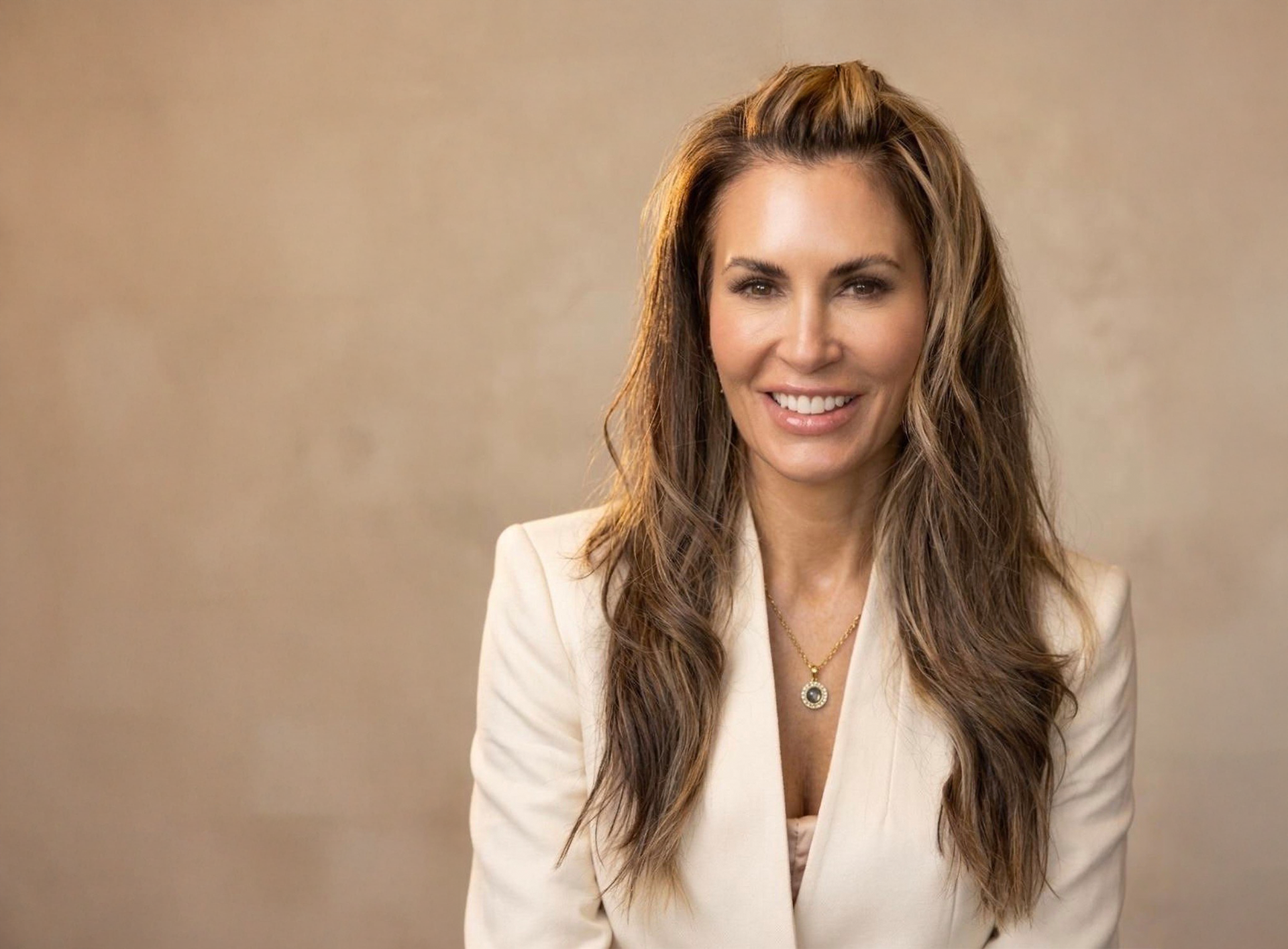 A smiling woman with long hair wearing a white blazer and pendant necklace against a soft, neutral background.