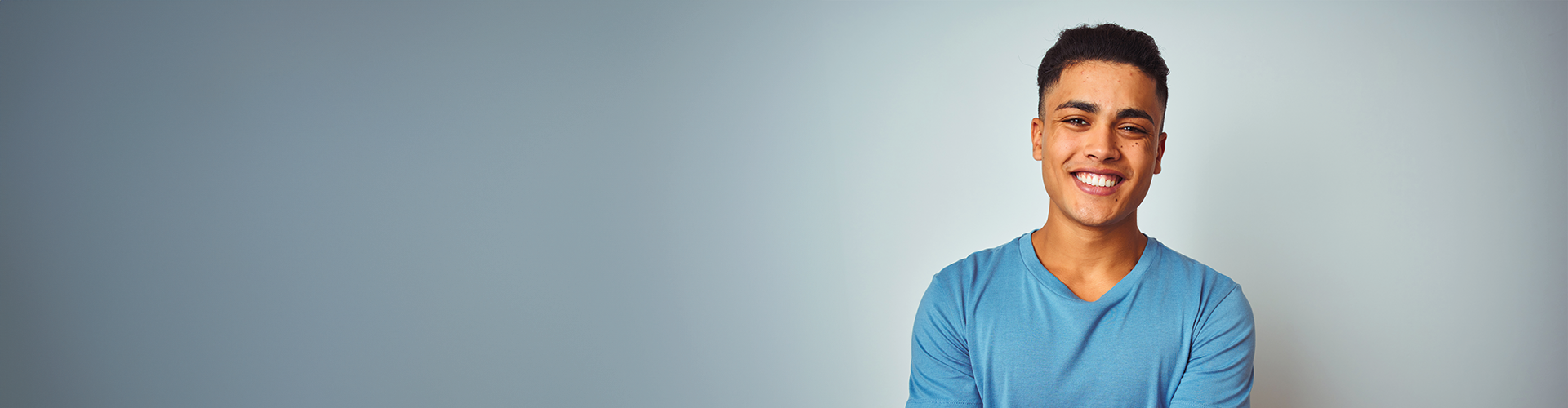 A smiling young man in a blue shirt stands against a light gray background.