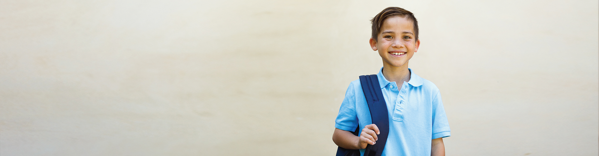 A smiling young boy wearing a light blue polo shirt holds a backpack over one shoulder against a neutral background.