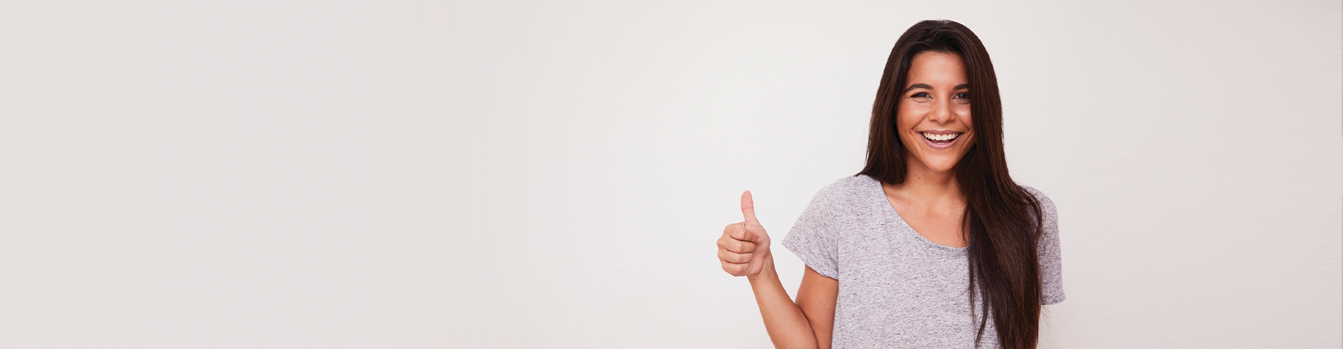 A young woman smiling and giving a thumbs up against a light-colored background, promoting a positive dental experience.