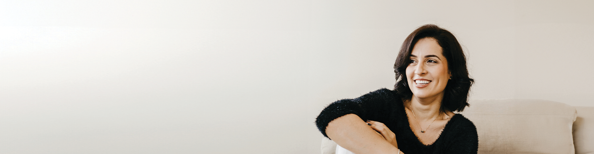 A woman smiling while seated on a couch against a plain background.