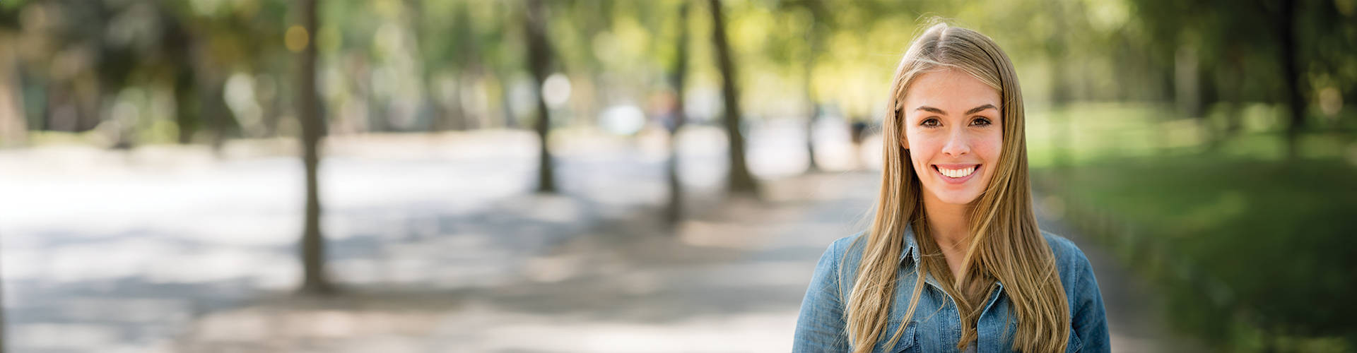 A young woman with long blonde hair smiling, standing on a tree-lined path in a sunny outdoor setting.