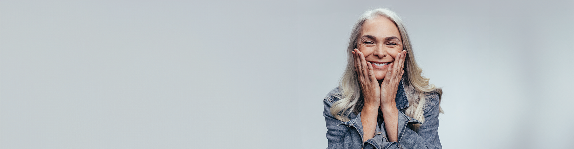 A smiling older woman with long gray hair holds her face with both hands against a light gray background.