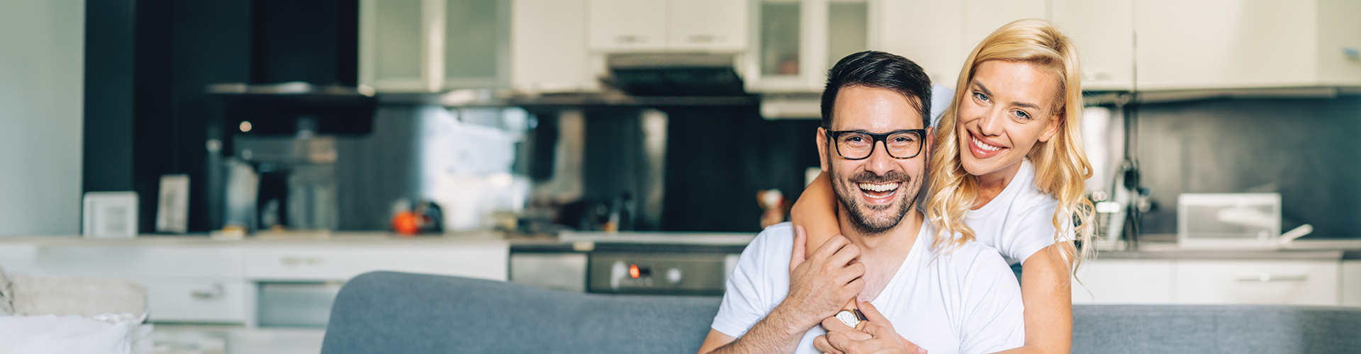 A smiling couple in casual clothes, embracing on a couch in a modern kitchen setting.