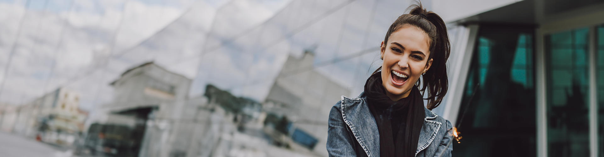 A smiling woman in a denim jacket stands against a modern, reflective building with cloudy skies in the background.