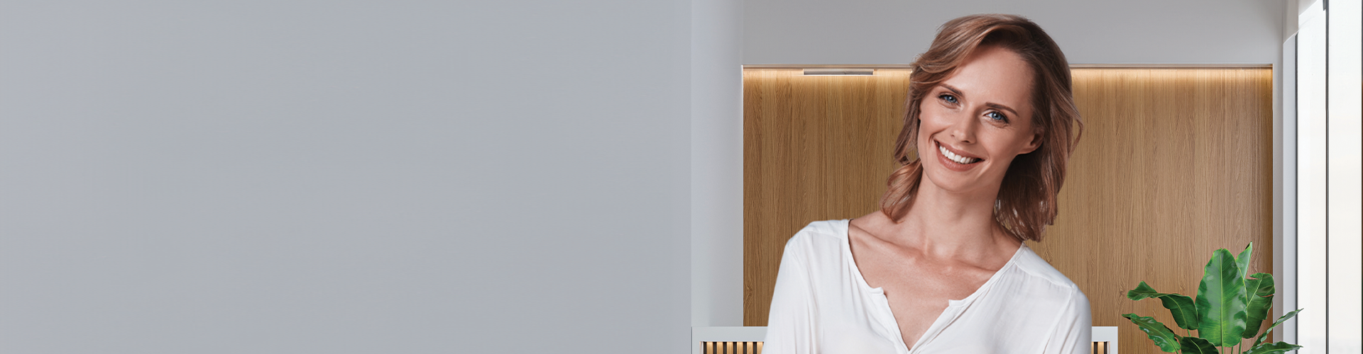 A smiling woman in a white shirt stands in a modern dental office with wooden accents and a plant nearby.