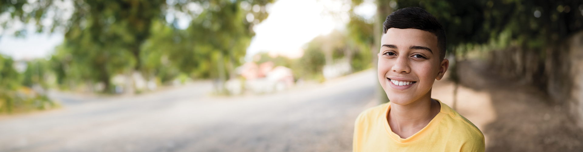 A young person wearing a yellow shirt smiles in a tree-lined outdoor setting with a blurred road in the background.