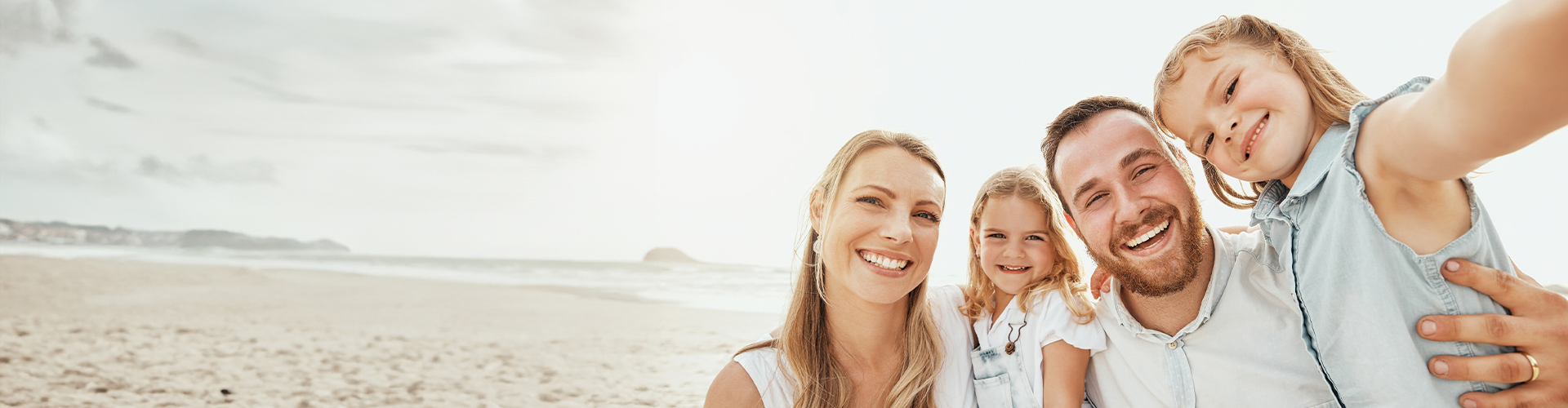 A family of four smiling for a selfie on a beach, with the ocean and distant coastline in the background.