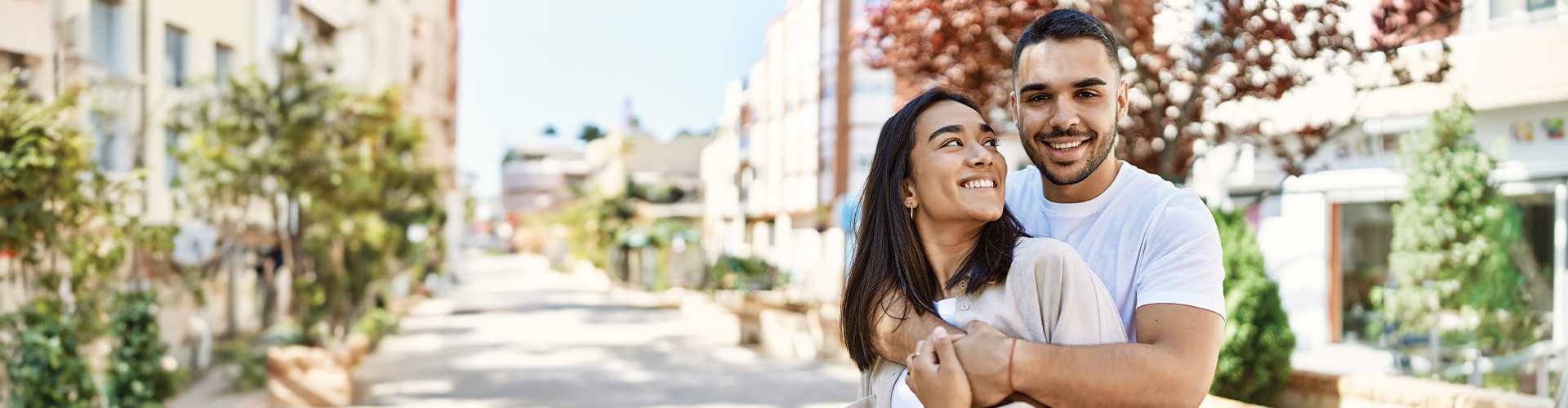 A couple smiling and embracing outdoors in a sunny, tree-lined street.