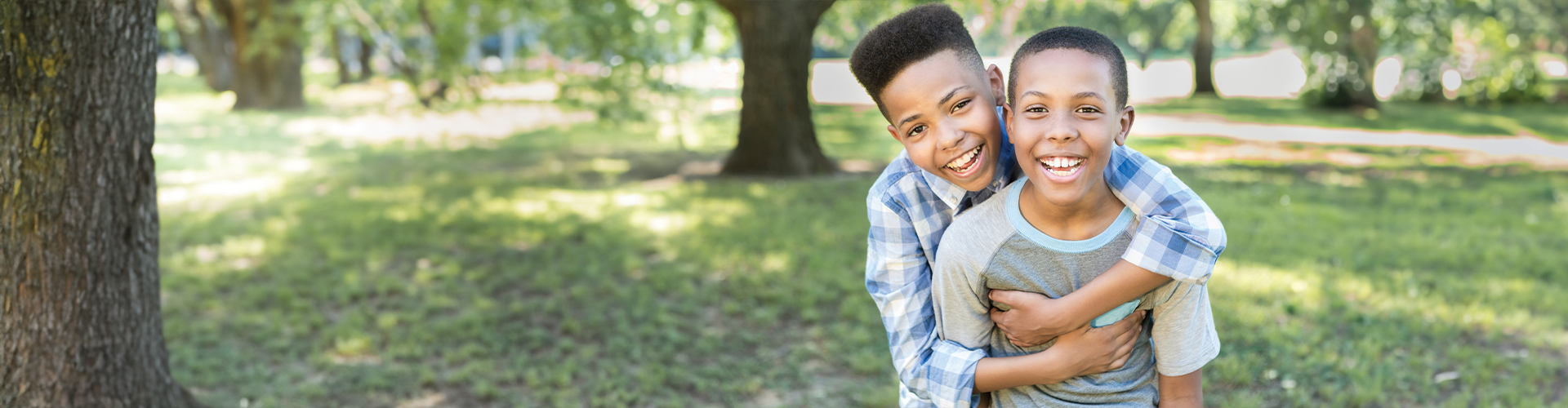Two young boys smiling and playing together outdoors, one boy giving the other a piggyback ride in a grassy park.