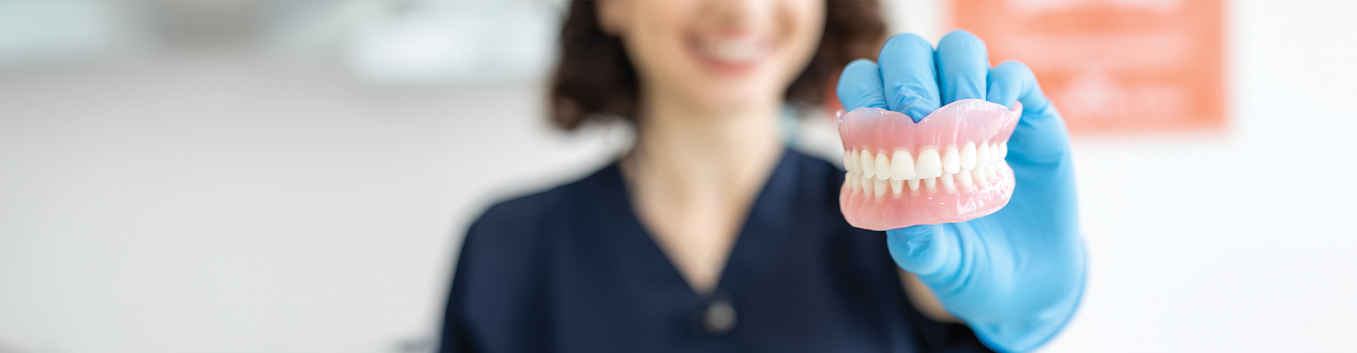 A dentist holding a dental prosthetic in gloved hands, smiling in a treatment room with soft lighting.