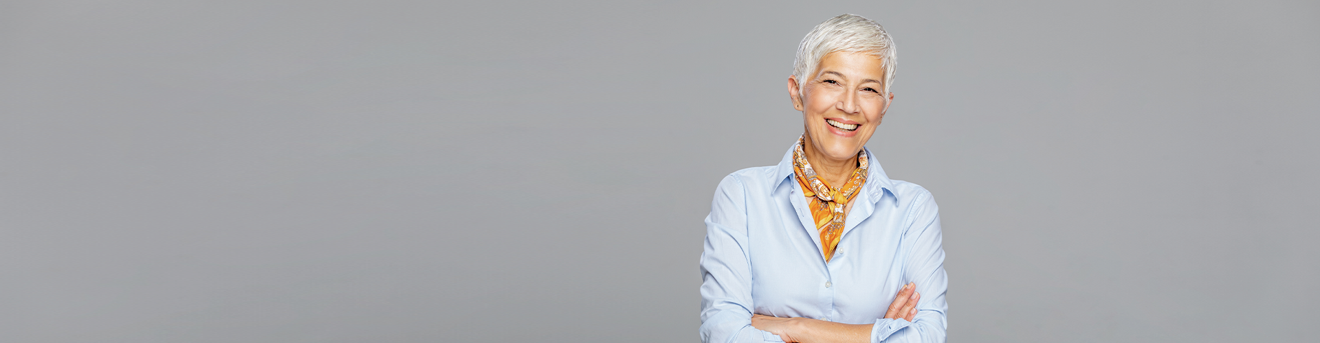 A smiling woman with short hair, wearing a light blue shirt and a patterned scarf, stands confidently with her arms crossed against a gray background.