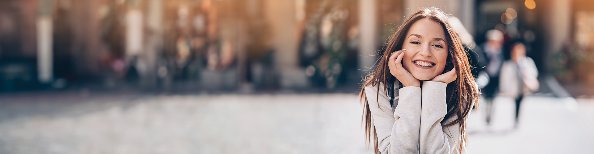 A young woman with braces smiles and poses happily outdoors, with a blurred background of people and buildings.