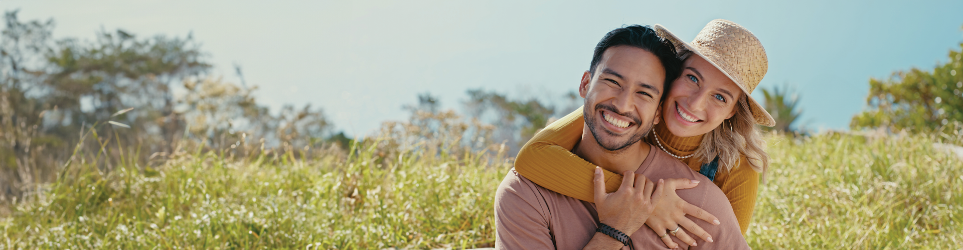 A smiling couple posing together outdoors, with the woman playfully hugging the man, surrounded by tall grass and trees.
