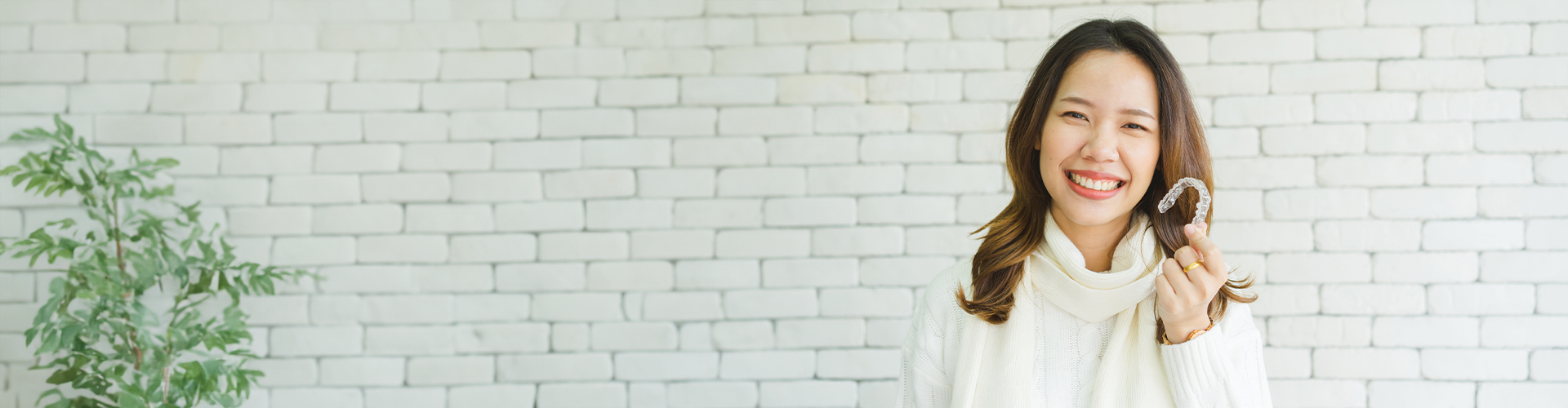 A woman in a white sweater smiles while holding a clear dental aligner in front of a white brick wall.