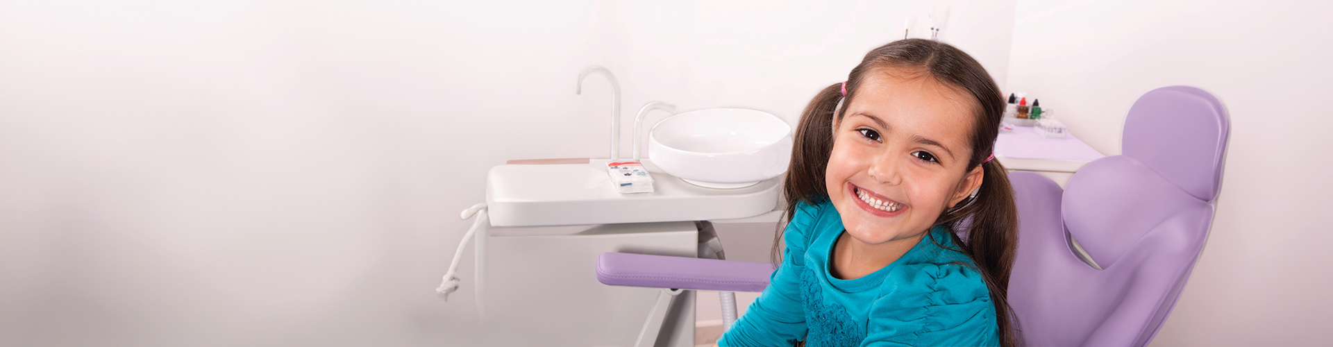 A smiling young girl sitting in a purple dental chair in a treatment room with a dental basin and equipment nearby.