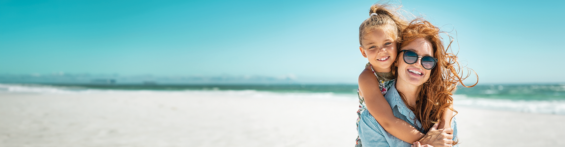 A woman and a girl embrace happily on a sunny beach, with turquoise water and a clear sky in the background.