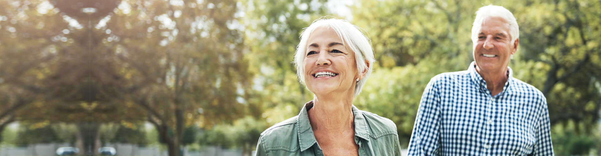 An older couple smiling and walking together outdoors among trees on a sunny day.