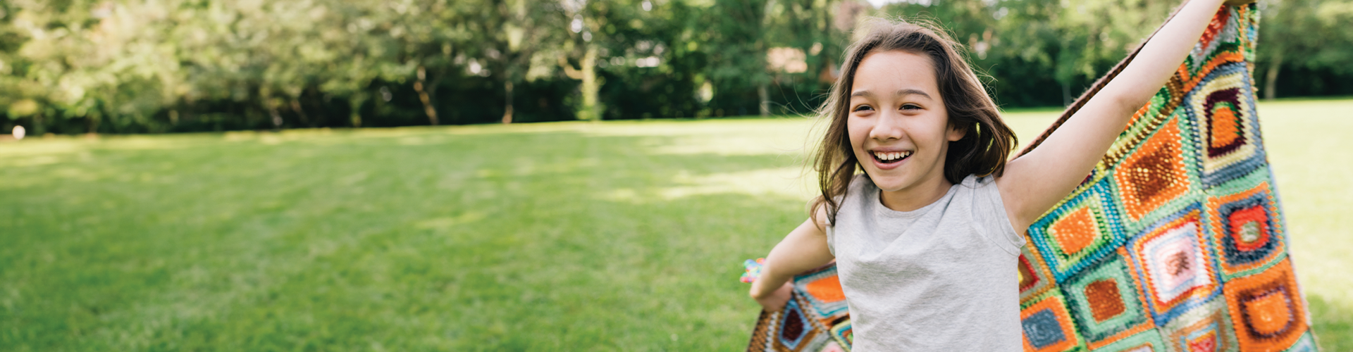 A girl joyfully runs across a grassy field while holding a colorful blanket.