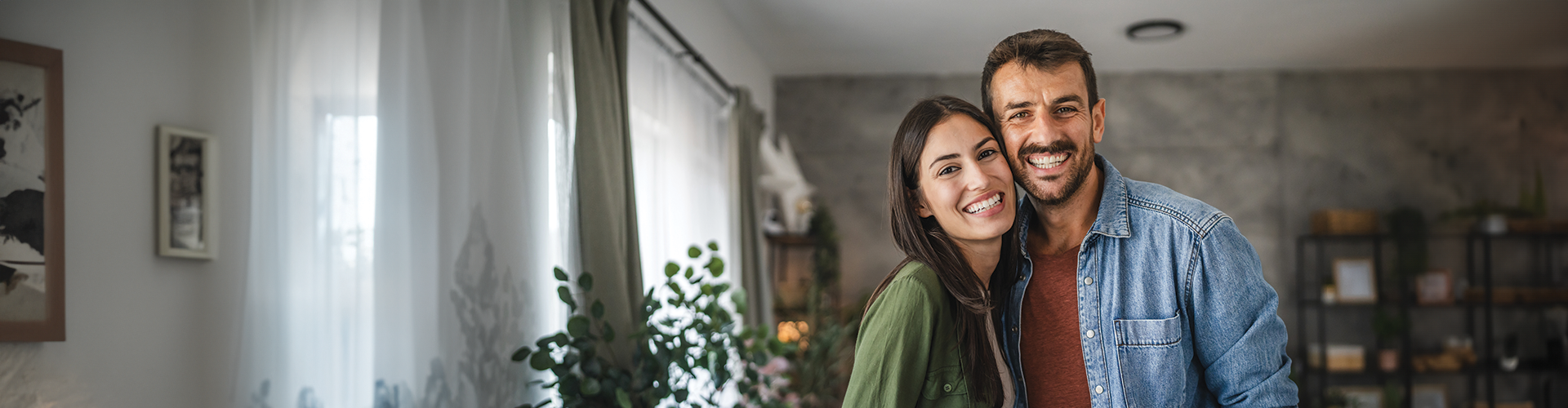 A smiling couple poses together in a cozy indoor setting with plants and artwork visible in the background.