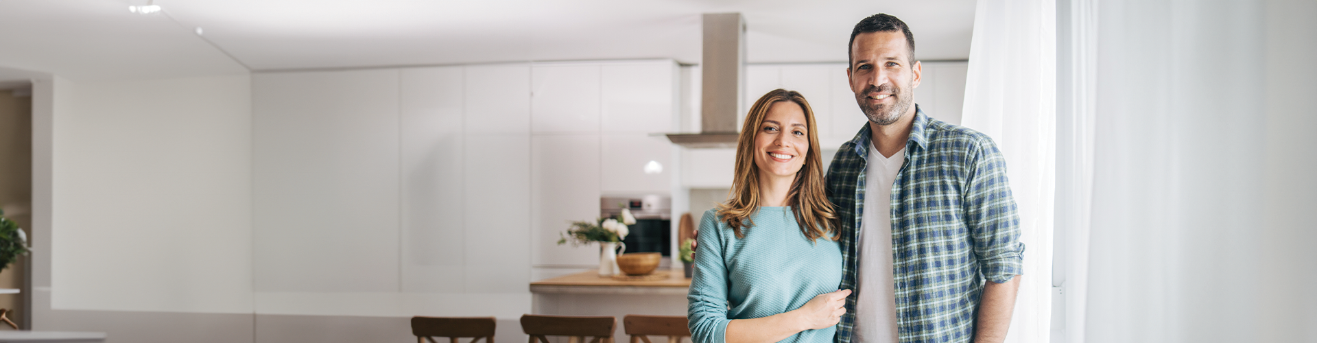 A smiling couple stands together in a modern kitchen with light walls and wooden accents.