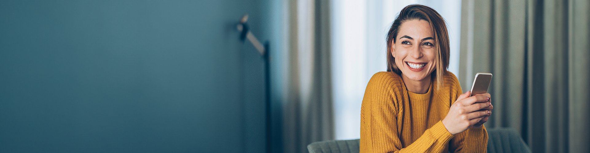 A smiling woman in a yellow sweater holds a smartphone while sitting in a cozy, well-lit room.