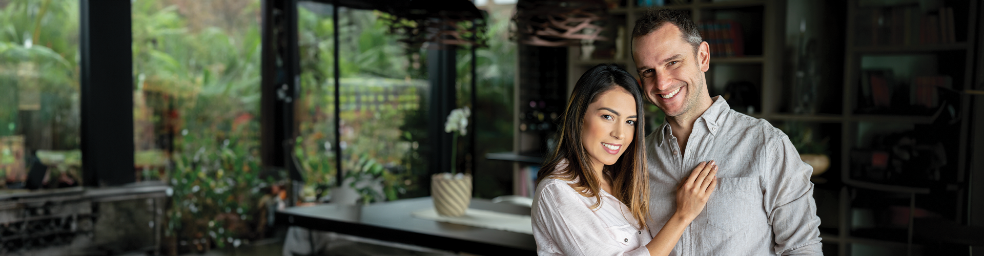 A woman and man smiling together in a modern, brightly lit room with greenery visible through large windows.