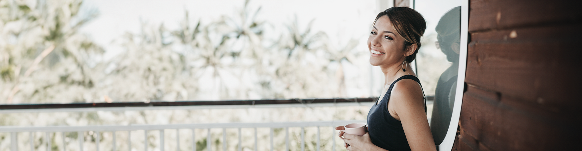 A woman smiling while holding a cup on a balcony with palm trees in the background.