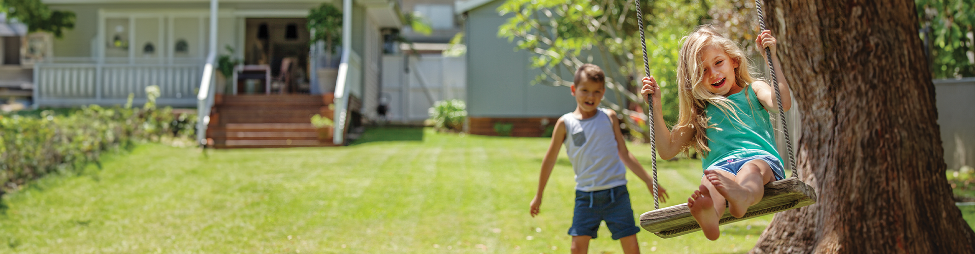A girl swings joyfully while a boy watches her from the backyard of a house on a sunny day.