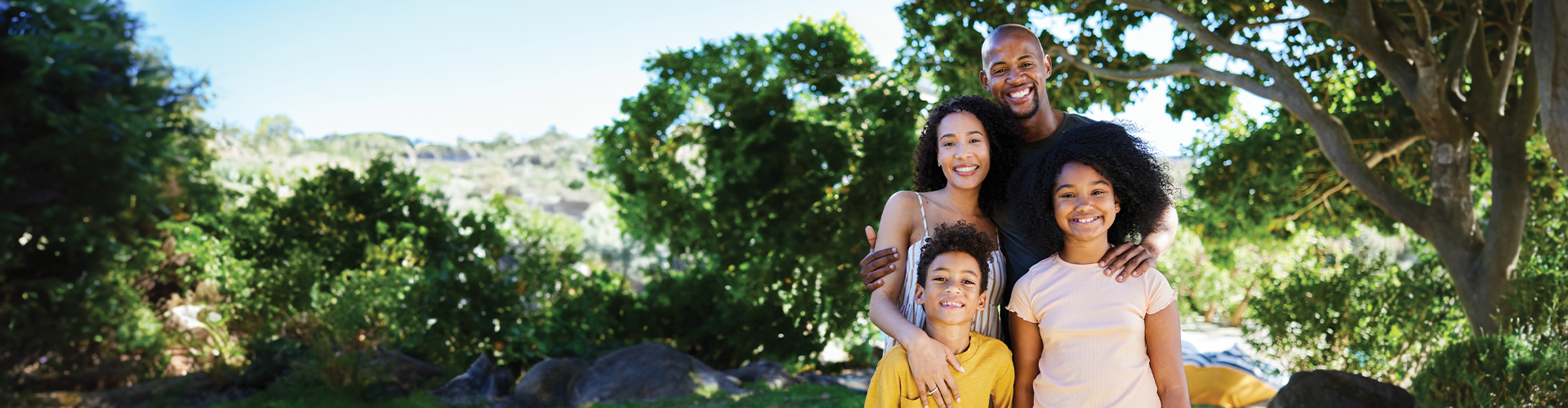 A smiling family of four stands together outdoors, surrounded by greenery and trees.