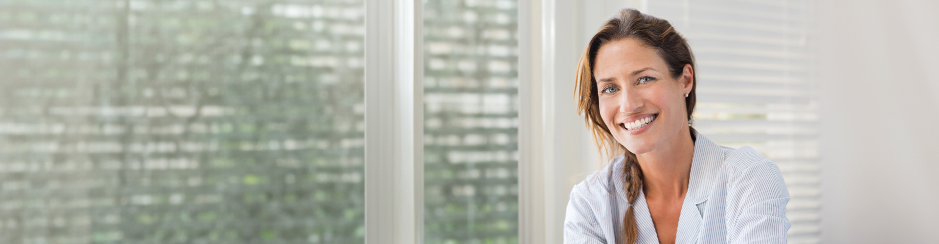 A smiling woman in a striped shirt sits at a desk with bright natural light and blinds in the background, suggesting a dental office.