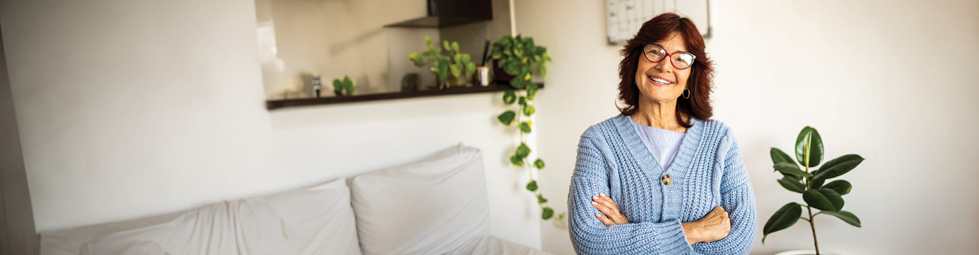A smiling older woman wearing glasses and a blue sweater stands with arms crossed in a bright, comfortable indoor setting.