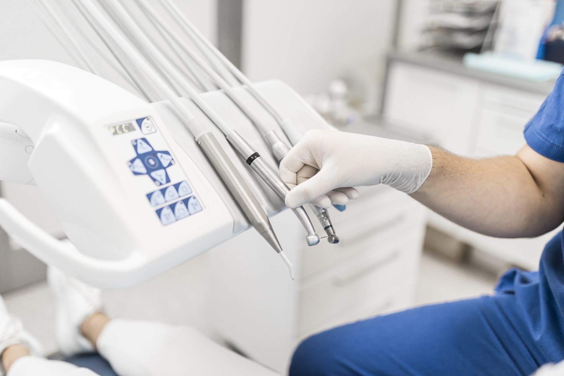 A dental professional in gloves selecting an instrument from a dental unit in a modern treatment room.