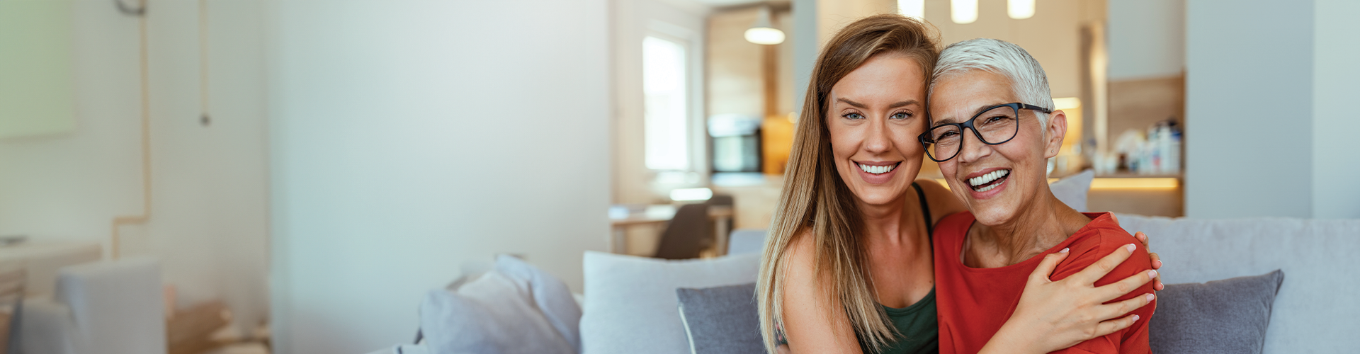 A young woman and an older woman smiling together on a couch in a bright, modern living space.