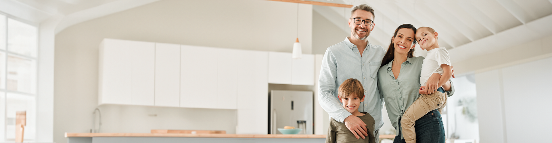 A smiling family of four stands together in a modern kitchen, with parents and two young boys, one being held by the mother.