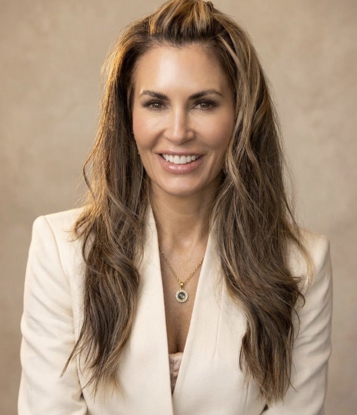 A smiling woman with long, wavy brown hair wearing a cream-colored blazer and necklace against a neutral background.