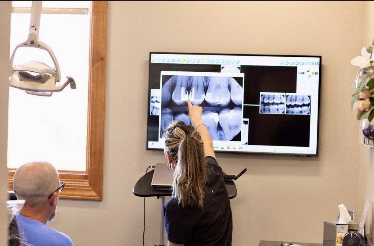 A dental professional pointing at dental X-rays on a screen while consulting with a patient in a dental office.