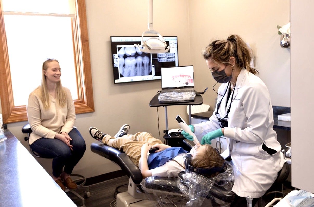 A dentist using a laser tool on a child in a dental treatment room while a parent observes.