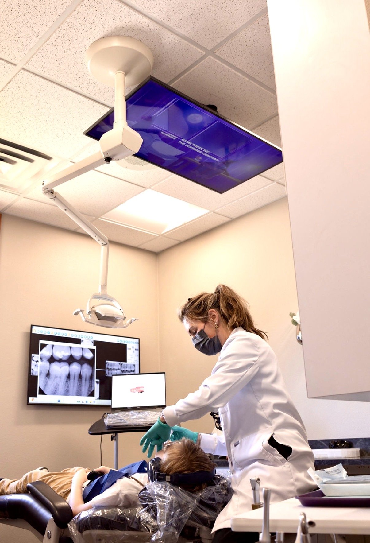 A dentist in a treatment room using laser dental methods on a child while examining dental images on digital screens.